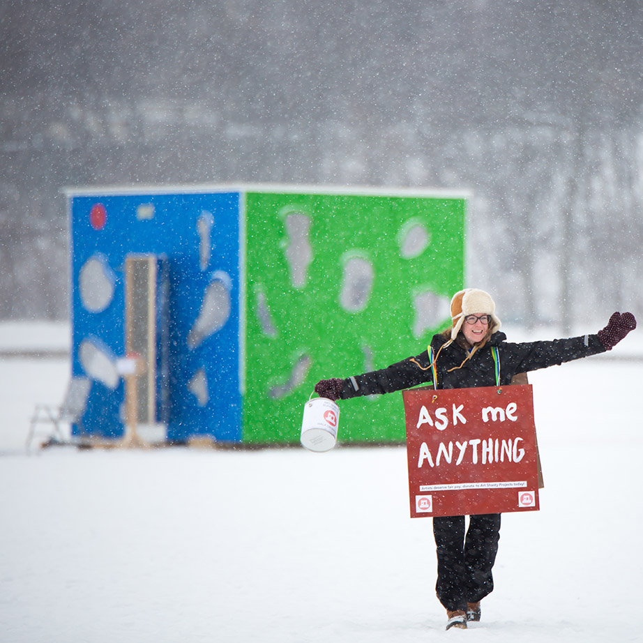 A woman walks away from a blue and green shanty, wearing a sign that says 'Ask me anything'