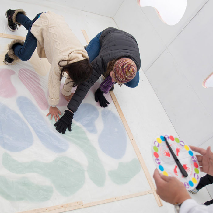 2 people crouched down playing the game twister, touching lake-shaped blobs, while hands hold a twister spinner.