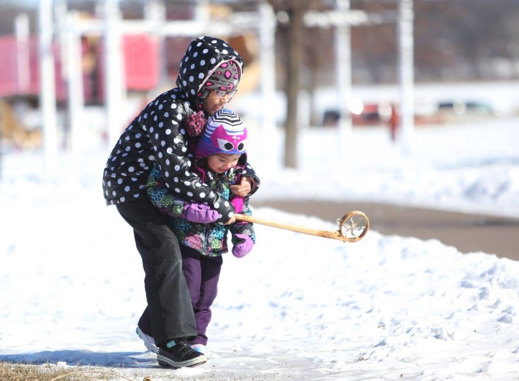 Two children on the snow with a lacrosse stick, the bigger one has their arms around the smaller one, and they both smile.