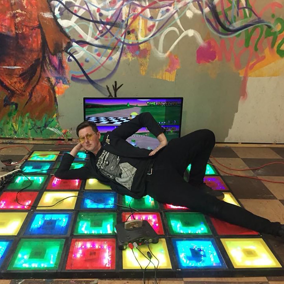 A person poses sassily, laying across a reactive, light-up disco dance floor. One hand is on their hip, the other is holding their head up. They're dressed all in black, and Mario Kart is playing on a big screen behind them.