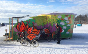 Outside the wildflower muraled Pollinator Shanty, a single person looks at the flock of monarch winged art bikes. The sky is light blue, with puffy white clouds.