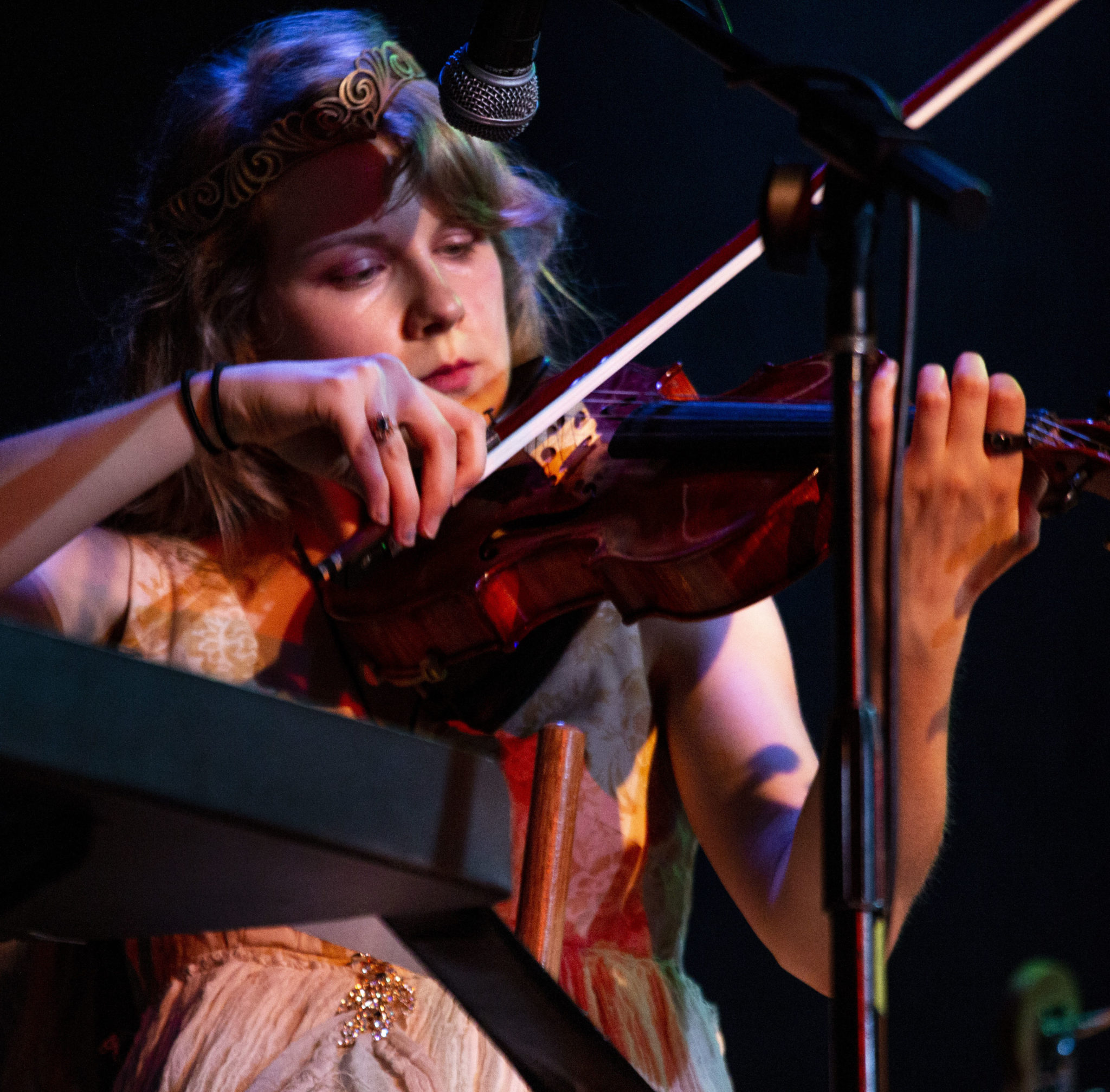A violinist, their eyes focused on the bow, pulls it across the strings near their chin. The shadows from different lights cast warm and cool colors across their face and arms.