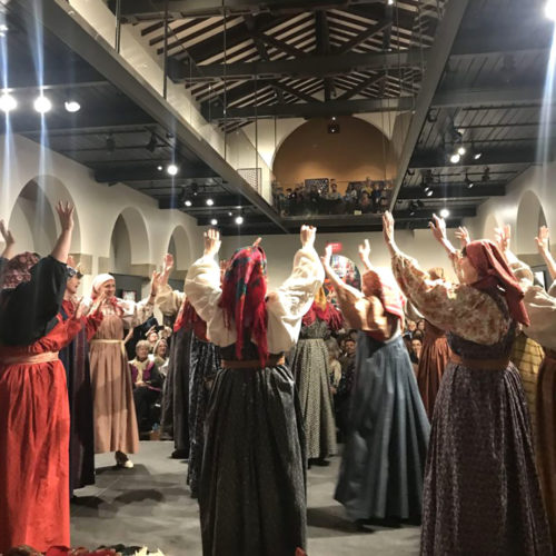 Many people in long dresses with full sleeves raise their heads up, dancing and maybe singing. They are all wearing headscarves. An audience watches from the museum's balcony above.
