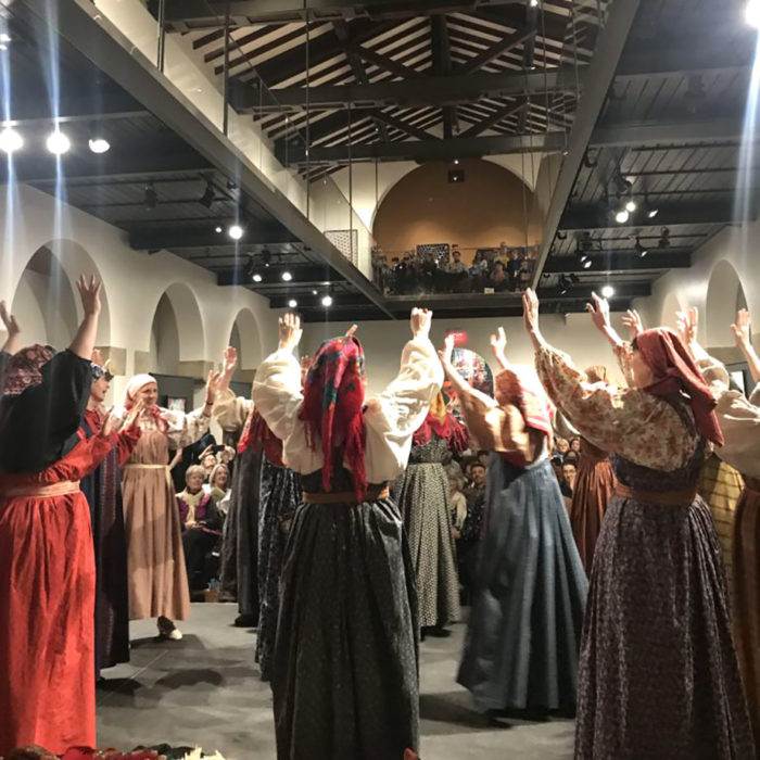 Many people in long dresses with full sleeves raise their heads up, dancing and maybe singing. They are all wearing headscarves. An audience watches from the museum's balcony above.