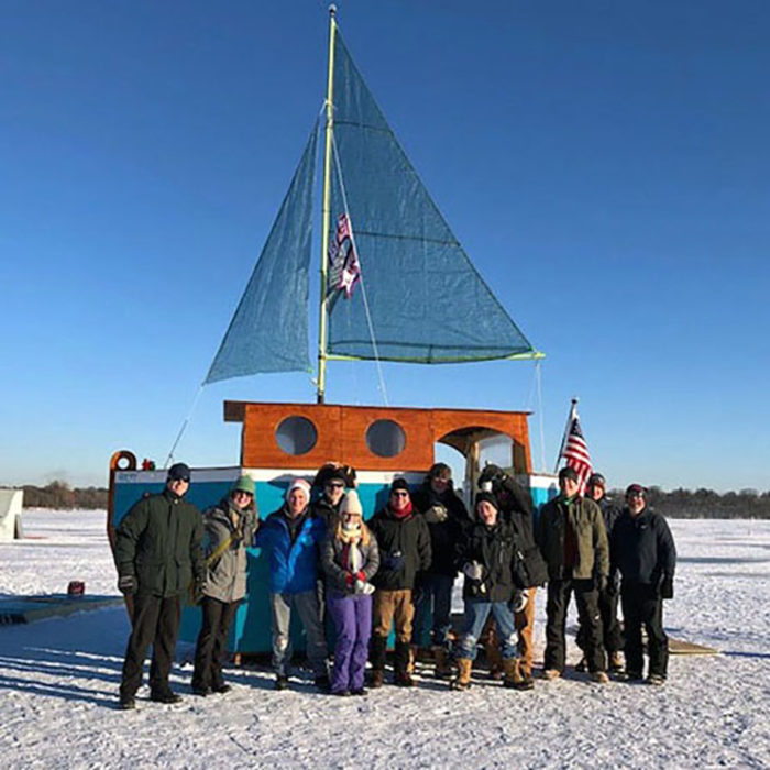 A dozen smiling people pose outside a sailboat on the ice.