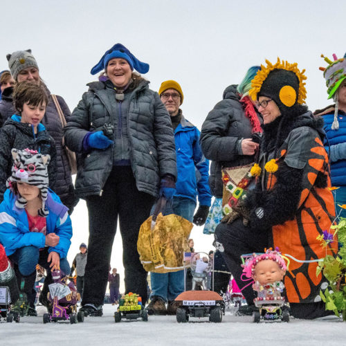 Smiling and laughing people crowded around tiny art cars on the snow- a child in a cat hat kneels, still dwarfing a purple car, the Mars rover, and a car with a babydoll head atop it.