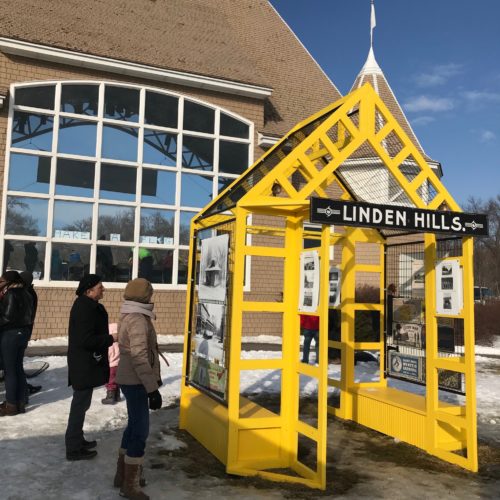 A few people stand, studying the yellow Linden Hills Neighborhood Shanty, in front of the Lake Harriet Bandshell. The yellow shanty is open and airy, and historic photographs of the neighborhood are hung on the open walls.