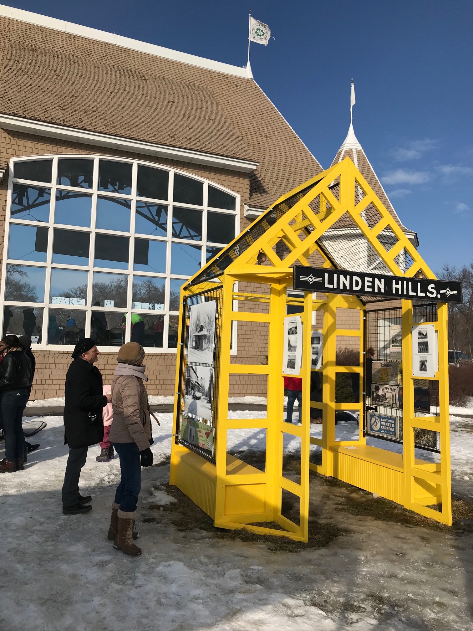 A few people stand, studying the yellow Linden Hills Neighborhood Shanty, in front of the Lake Harriet Bandshell. The yellow shanty is open and airy, and historic photographs of the neighborhood are hung on the open walls.