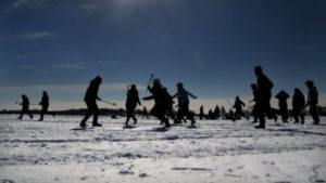 A dozen silhouettes of lacrosse players caught in dynamic motion on the snow- two players run towards the left edge of the photograph, four seem to converge in the center, two with sticks raised. More players or perhaps observers look in from the right.