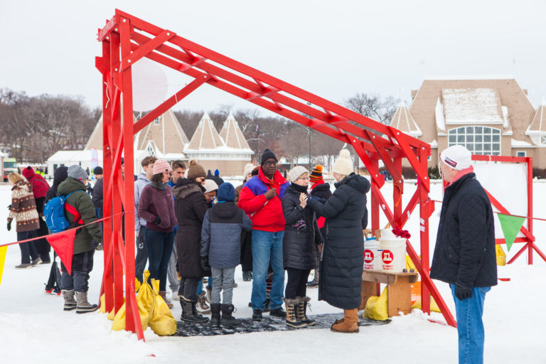 A volunteer attaches a ski tag to a new shanty villager. A thick crowd of people wait to enter through the bright red shanty welcome gate.