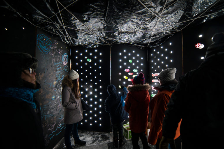 Inside a dark shanty, backlit forms of children and adults in winter gear reach towards holes in the black walls where light streams in from outside.
