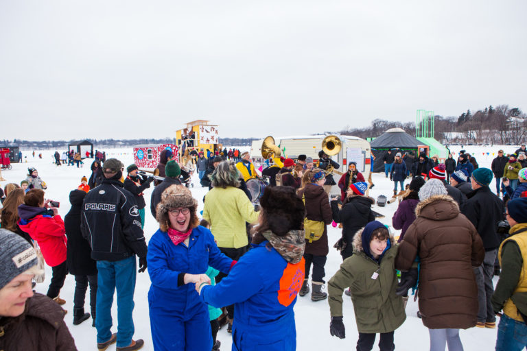 Bundled up artists and audiences joyfully dance to the tunes of Brass Messengers during a pre-pandemic day at the colorful shanty village on frozen Bde Unma in Minneapolis, February 2020.