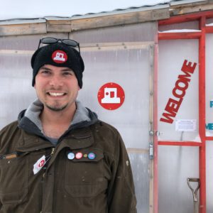 Smiling man stands in front of the Welcome Shanty with snow falling