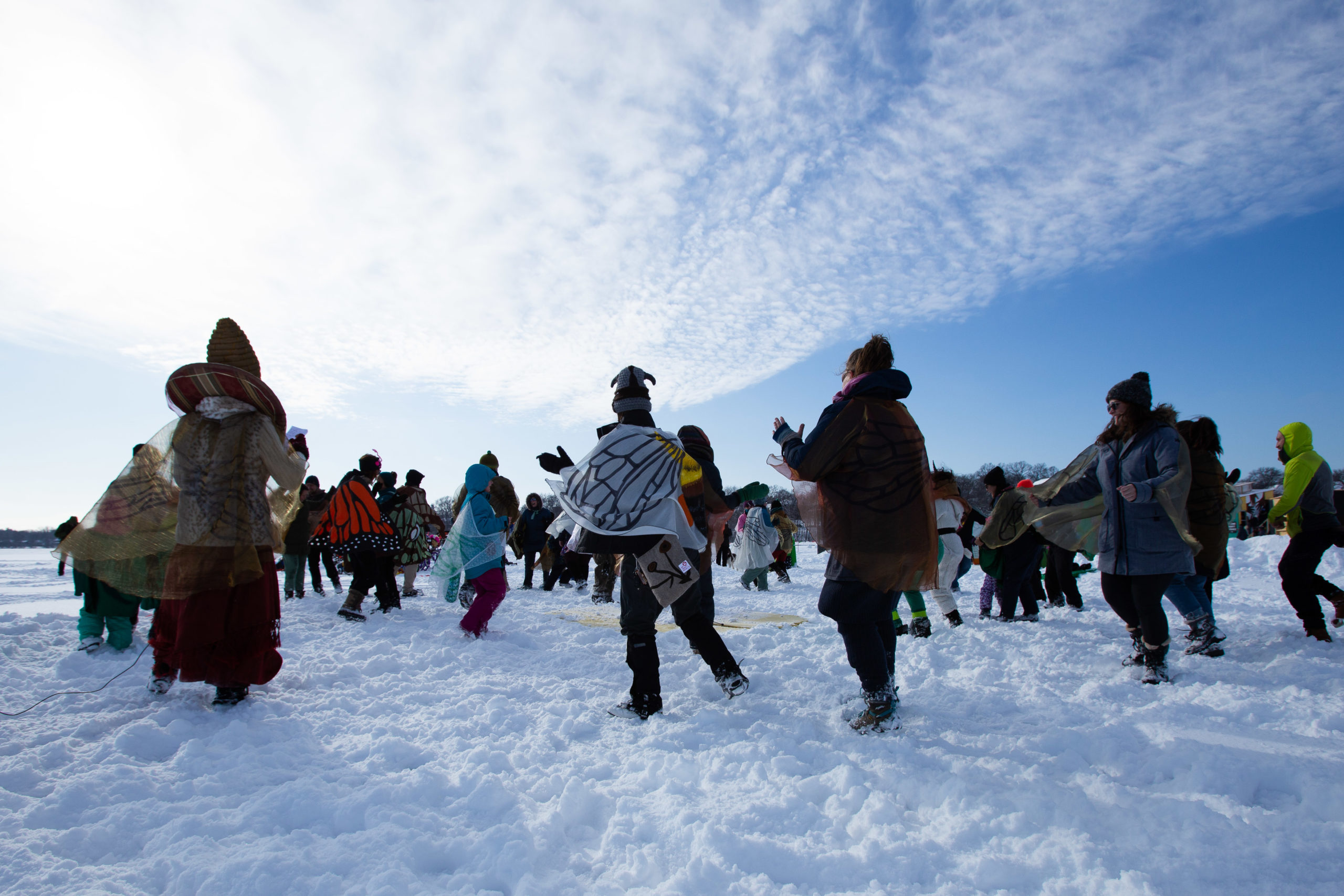 A group of adults dance in the trampled snow, in a circular formation, facing the center. Some wear butterfly wings on their bundled up bodies.