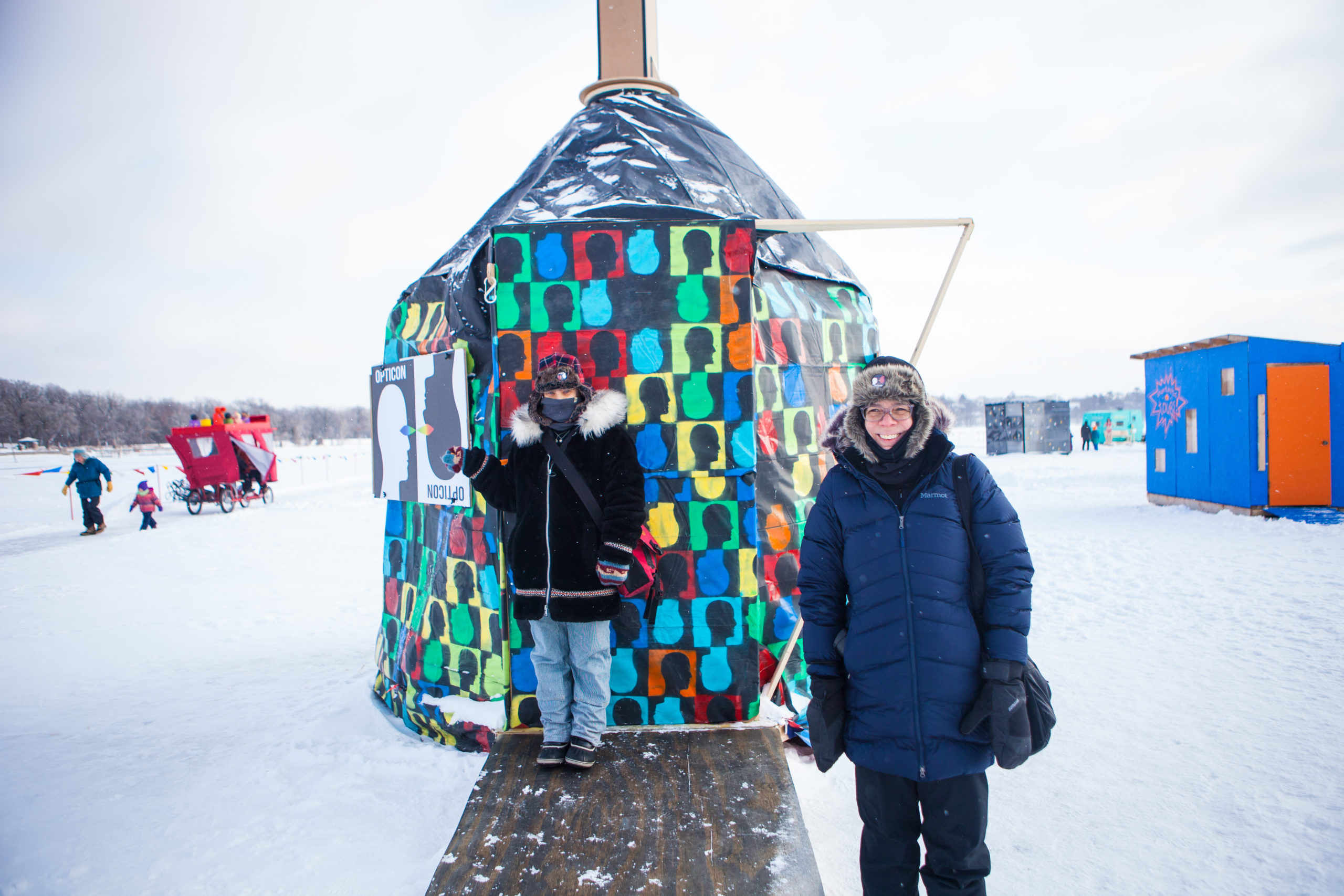 An bunded up artist stands in front of a colorful yurt on a frozen lake. Other shanties and people are seen in the distance, across the ice.