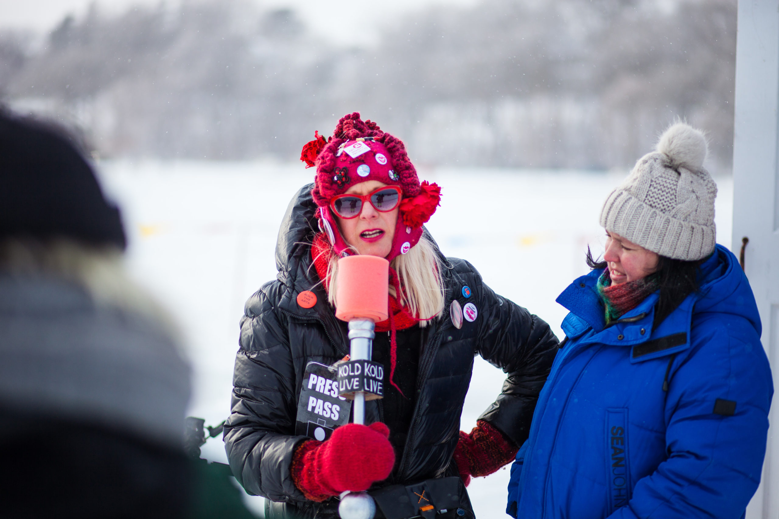 Bundled up and wearing red sunglasses, Patty Pucker holds an oversized prop microphone, brow furrowed slightly and one arm on her hip. A person in a blue parka standing next to her listens thoughtfully.