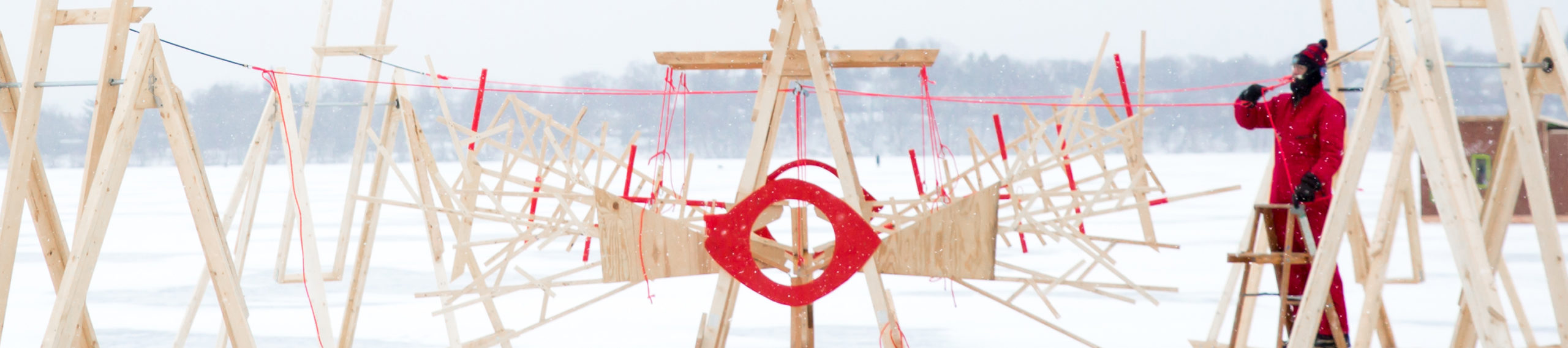 An adult stands on a ladder, working on a bright pink rope attached to a set of unpainted wooden structures. In the center structure, a huge, almond-shaped red eye is at the center of a sparse wooden pyramid.