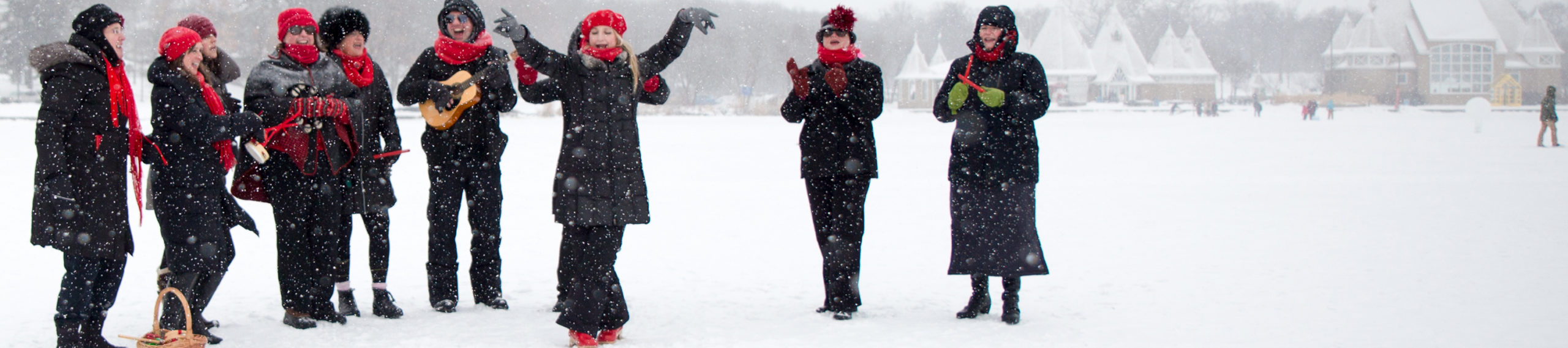 A joyful person in a red beret lifts their arms and sings. They are surrounded by people with red winter accessories with small percussion instruments and a small guitar, all singing merrily.