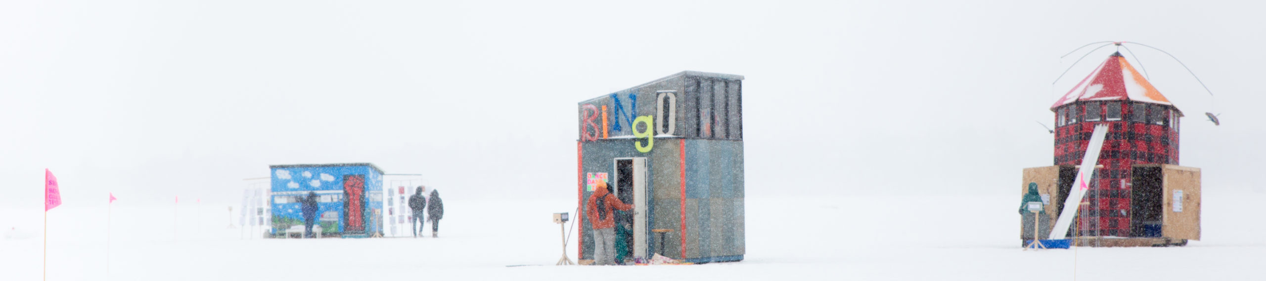 Almost pixelated by the snow, three shanties stand out against the white day: a small light blue shanty with a red door, two figures enter the bingo shanty, and a red plaid shanty that looks like a lighthouse.