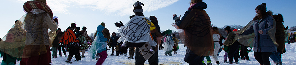 A group of adults dance in the trampled snow, in a circular formation, facing the center. Some wear butterfly wings on their bundled up bodies.