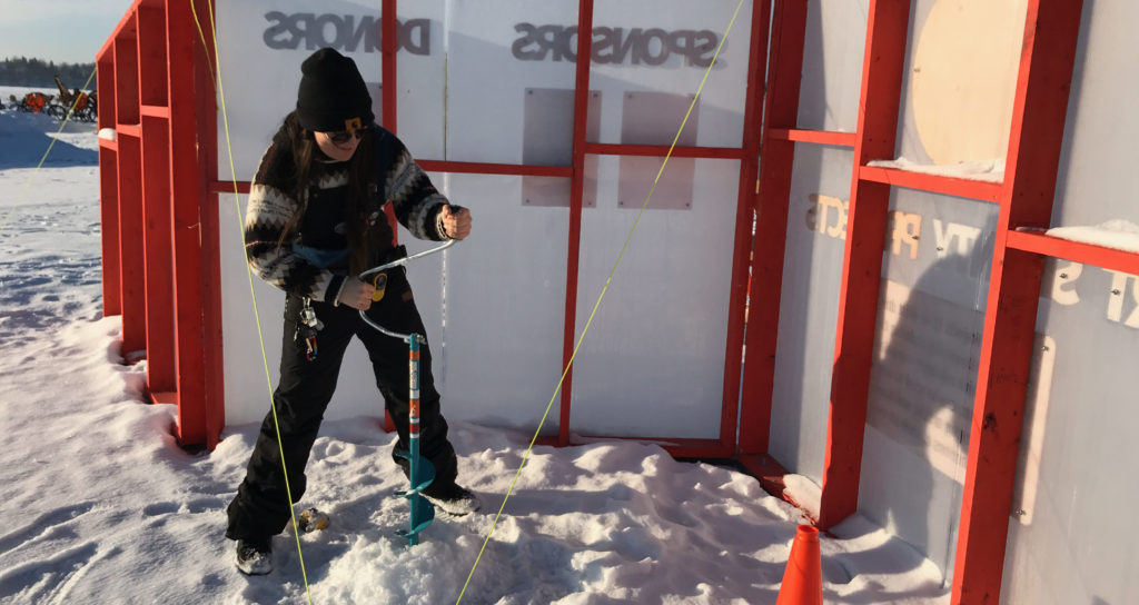 A staff member uses a hand crank auger to drill a hole in the ice behind a red and white information board on the frozen lake. 