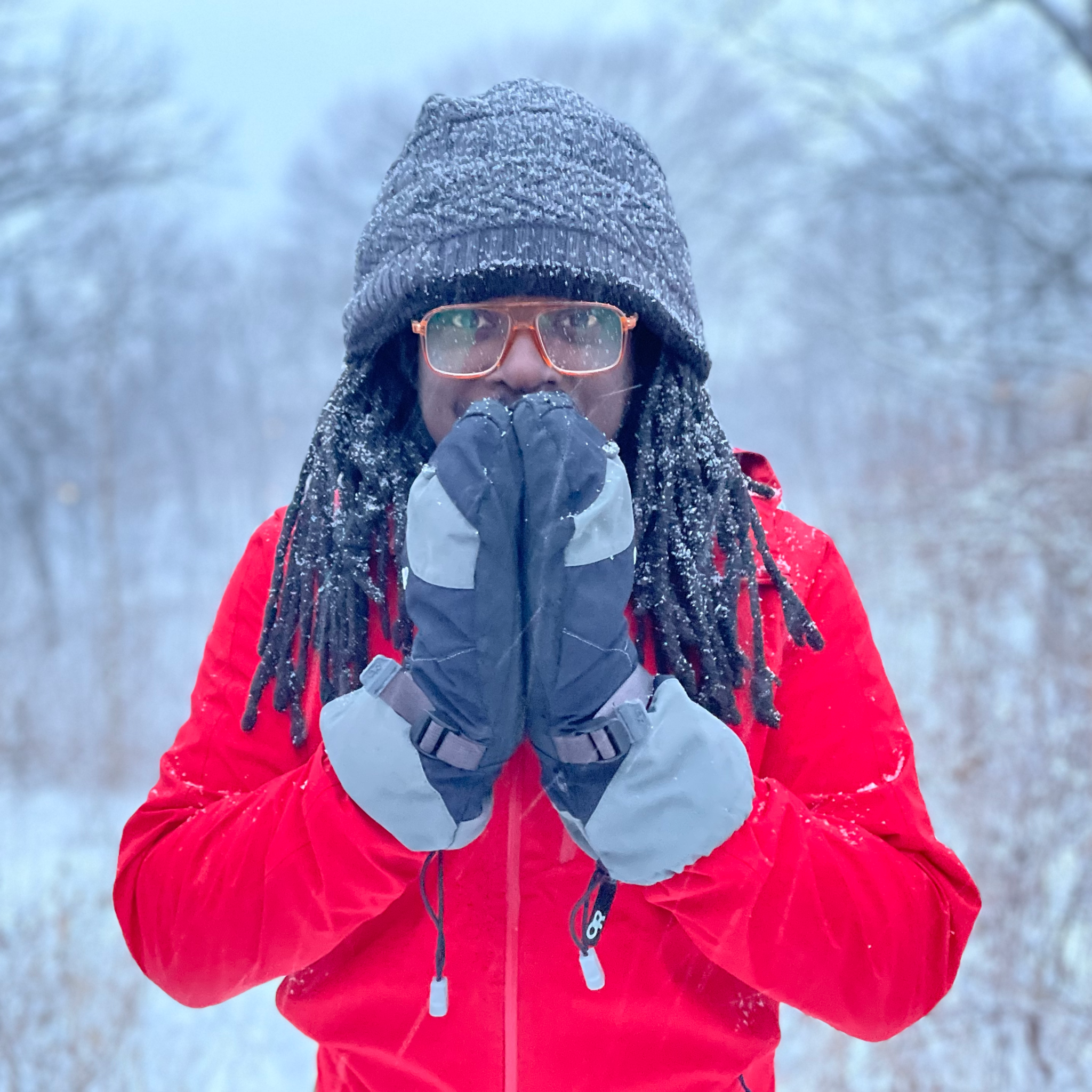 A person with dark skin and long locs stands under falling snow with their hands in prayer position.