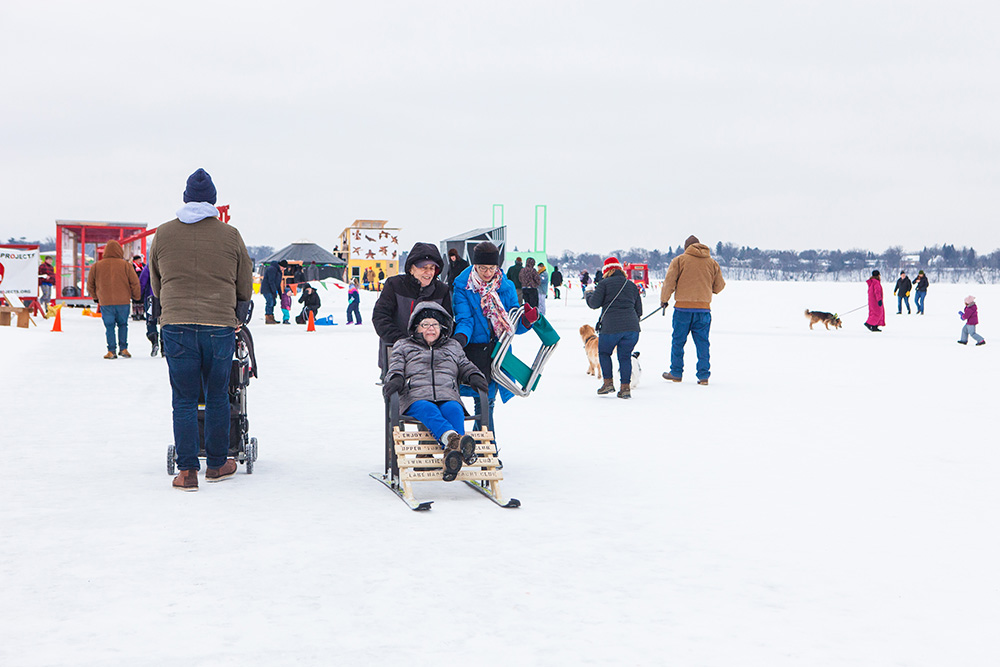 A visitor pushes their friend on a makeshift sled. There is a crowd of several people in the background.
