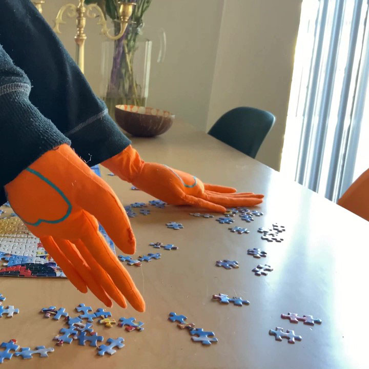 Arms with orange soft scupture hands reach towards puzzle pieces on a table