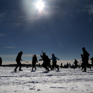A group of people play lacrosse in silhouette on a frozen lake.