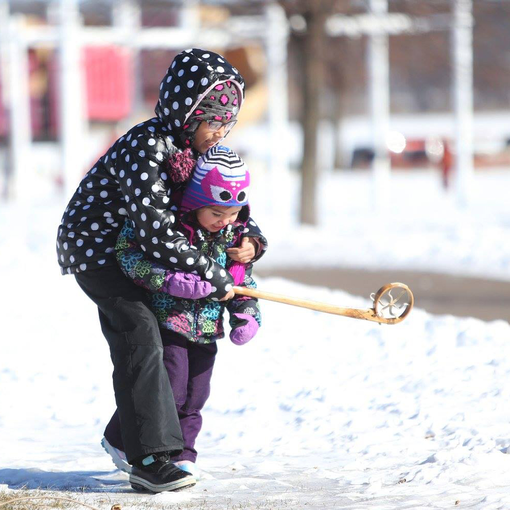 A school-aged kid helps a toddler with a wooden lacrosse stick. They play on a snowy area.