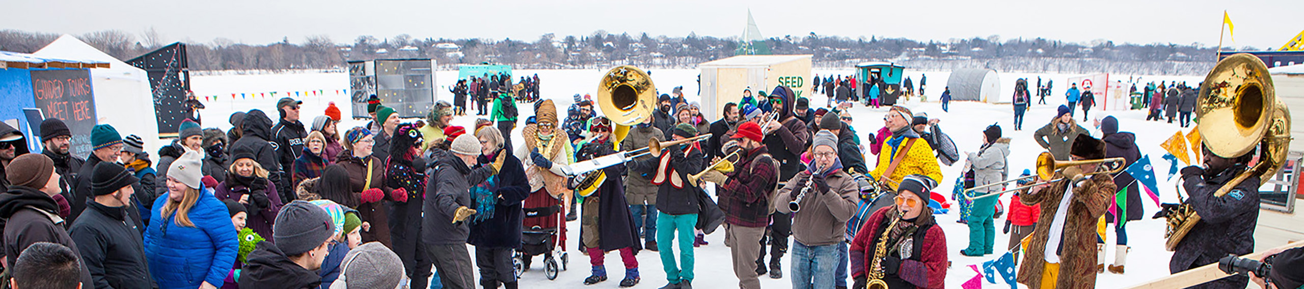 a large crowd in colorful winter-wear gathers around musicians on the frozen lake, with shanties and people in the background