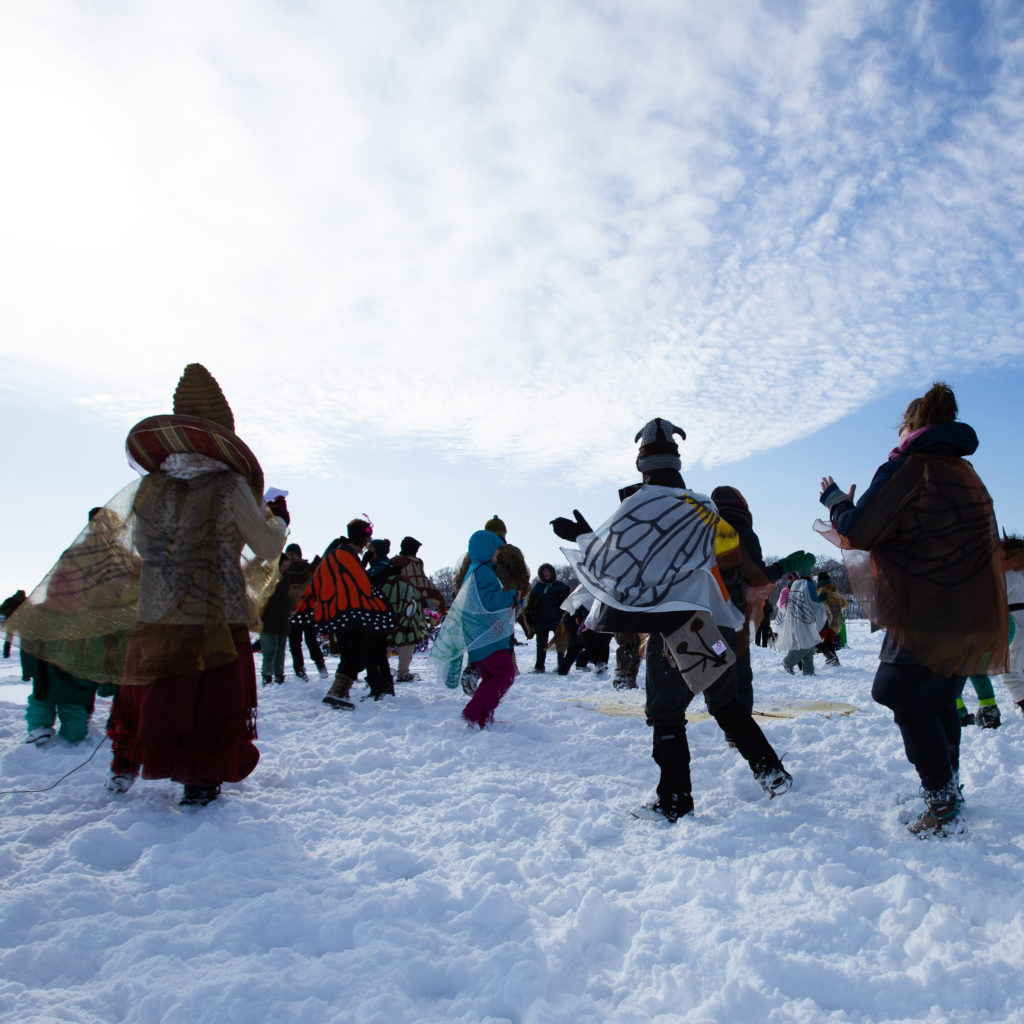 A group of adults dance in the trampled snow, in a circular formation, facing the center. Some wear butterfly wings on their bundled up bodies.