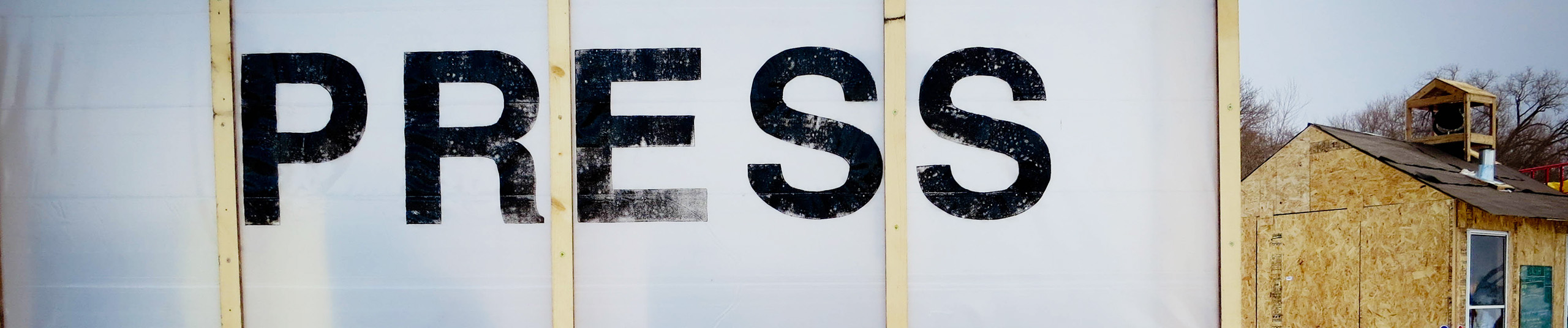 the letters "PRESS" painted in black on plastic wall, with a plywood shanty in the distance