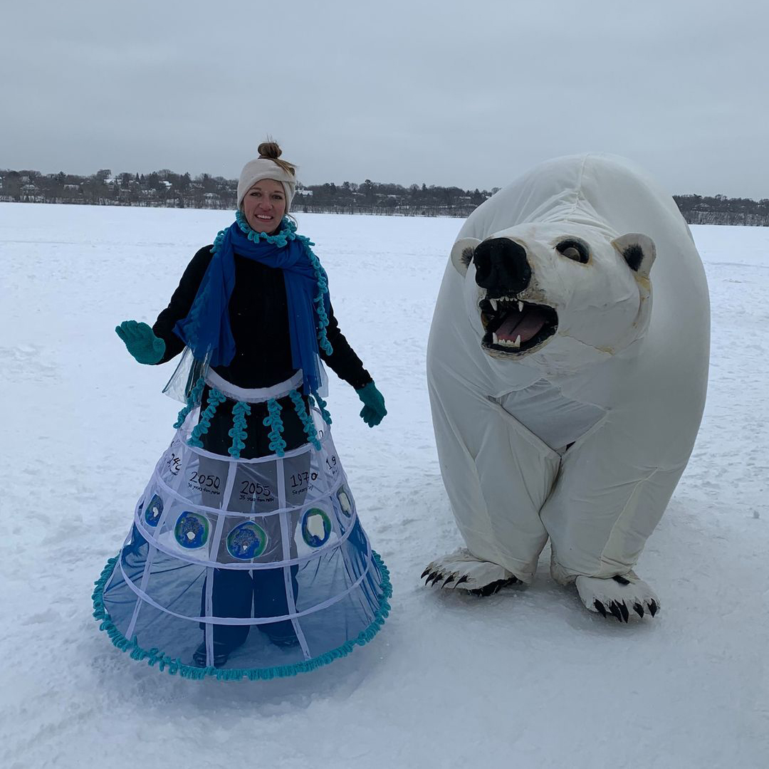 A person in an icy blue costume guides a giant polar bear puppet across a frozen lake