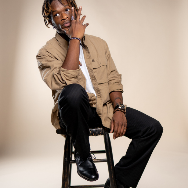 A man with locs casually sits on a stool and looks off to the side.