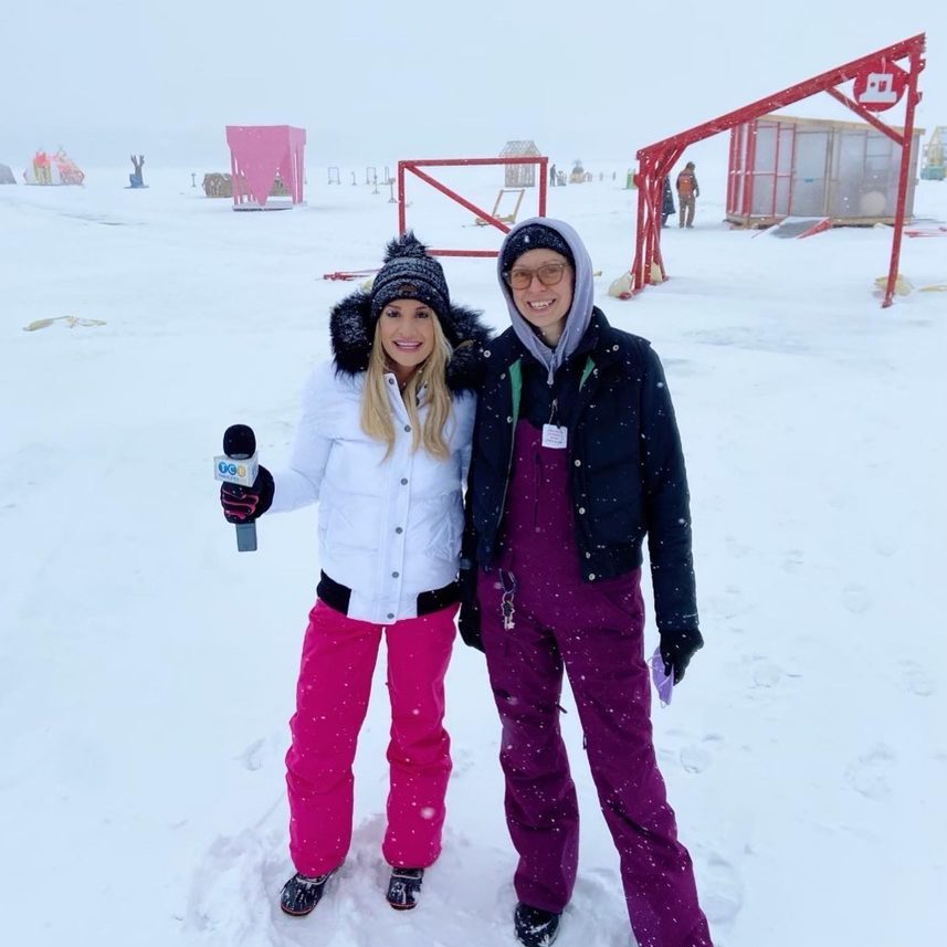 a TV host holding a mic poses with a staff member on the frozen lake. Both wear pink and purple snowpants.