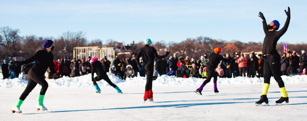 A group of ice skaters wearing black costumes and colorful berets dance for a crowd