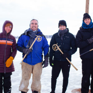 Four people pose holding traditional wooden lacrosse sticks