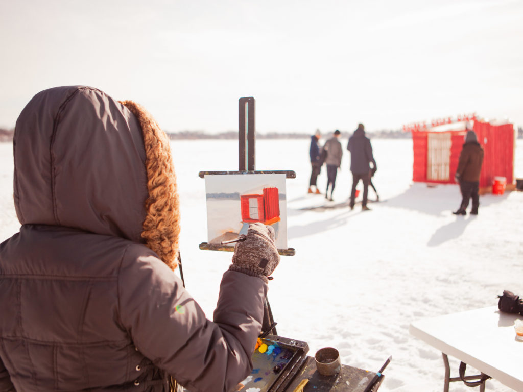 A bundled up person stands at an easel and paints a portrait of a shanty in the distance.