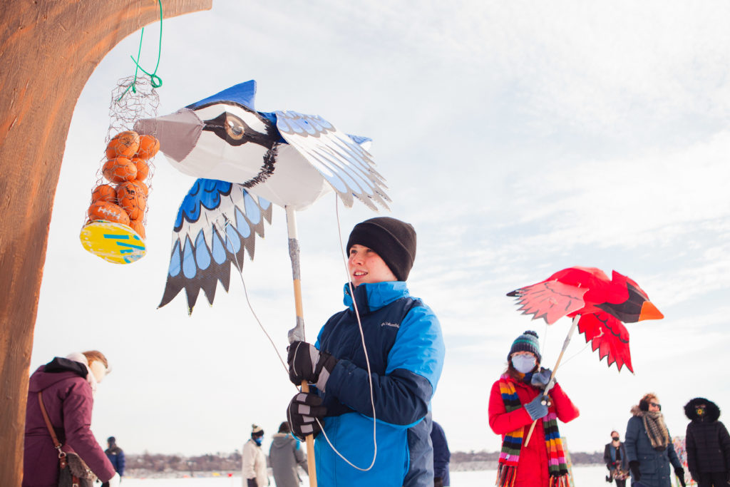 Two kids play with big vibrant bird puppets against a cloudy sky