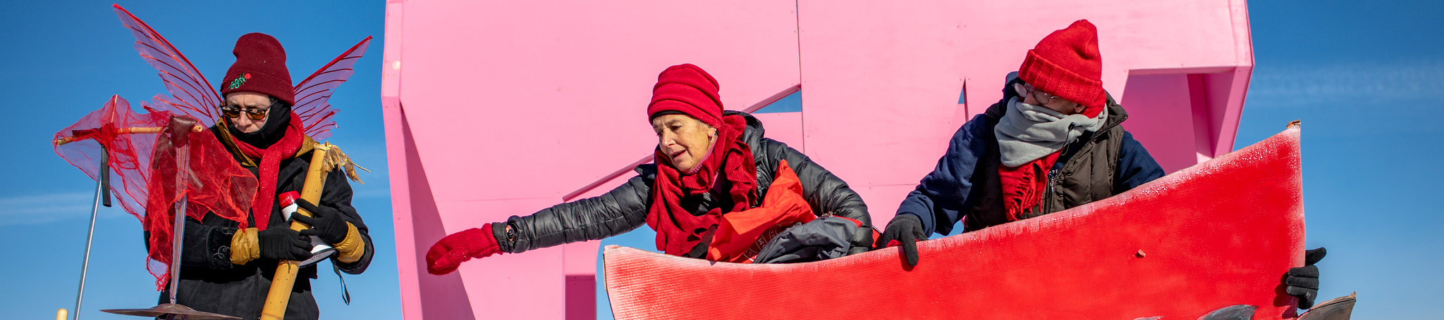 A theatre troupe in red costumes rows a red boat across a blue and pink background outside.