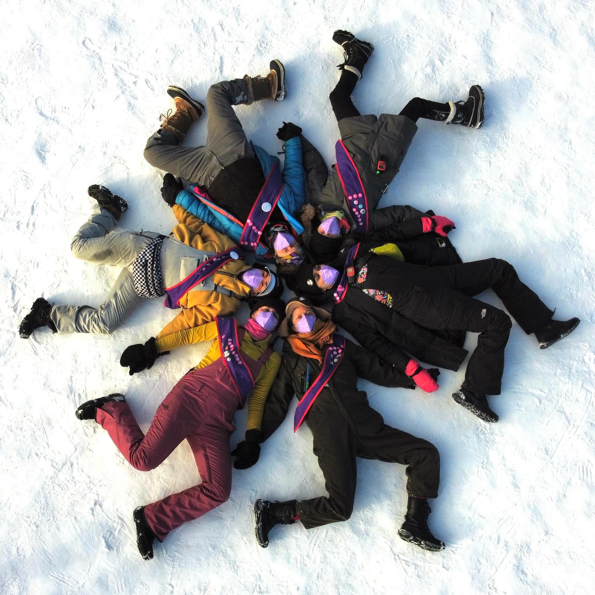 A view from above: six people in snowsuits lay with their heads in the center and feet extending out, in snowflake formation.