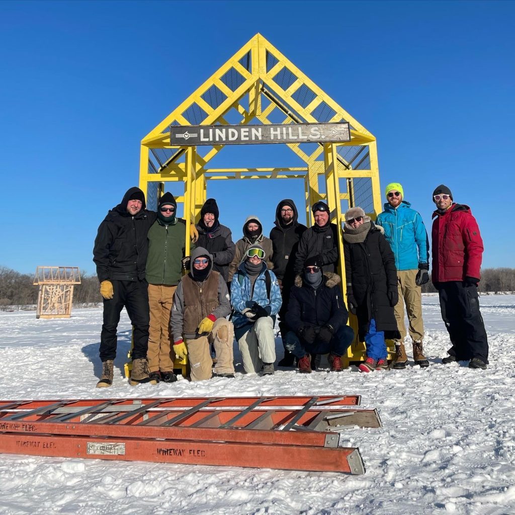 A group of people pose in front of a yellow arched shanty. A giant orange ladder lays on the snow in front of them.