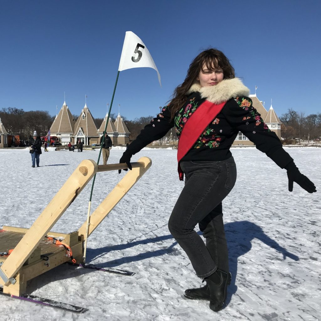 A person wearing a snowsuit and red sash poses while pushing a kicksled.