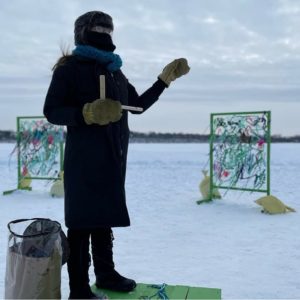 a very bundled up person stands on a platform and gestures with their hand to indicate the vibrantly woven shanty panels behind them.