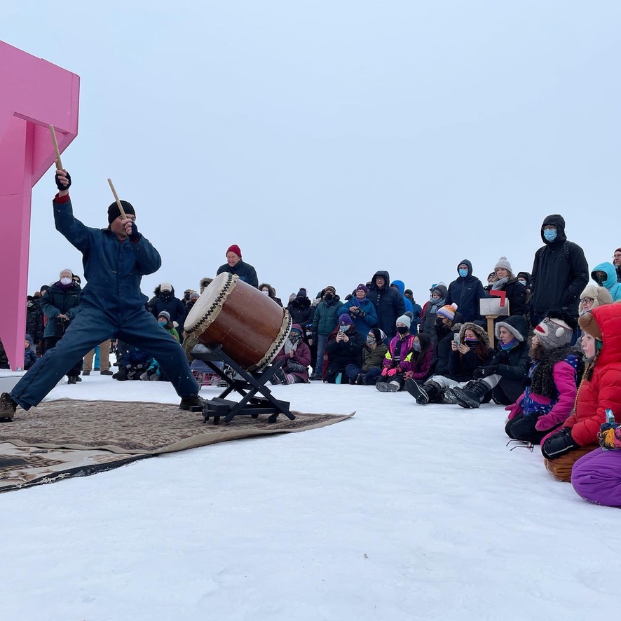 A person plays a taiko drum in front of a crowd that sits on the snowy frozen lake