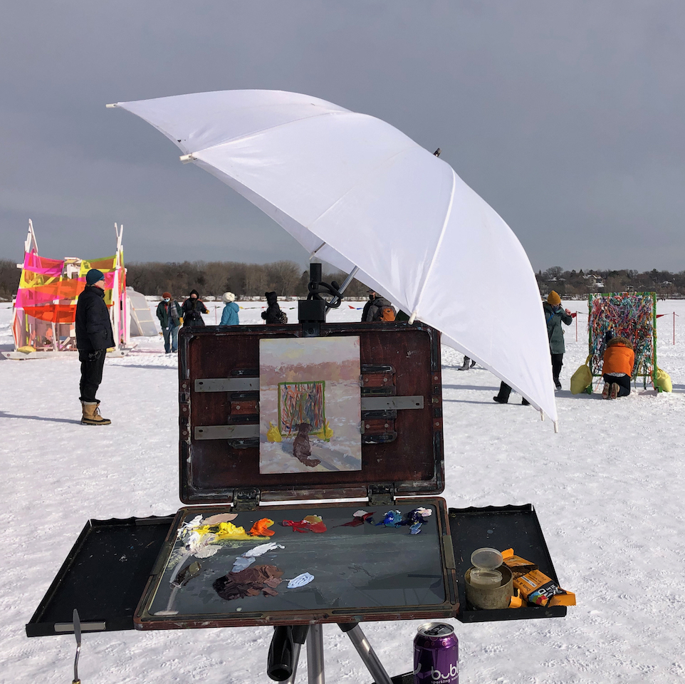 A plein air painting station is set up on a frozen lake with the shanty being painted in the distance.