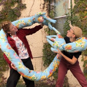 Two people hold a large soft sculpture around them, as if hugging.