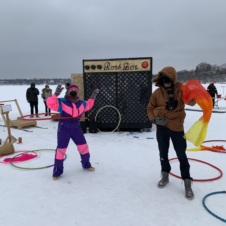 People in snowsuits hula hoop in front of an amp-shaped shanty on a frozen lake.