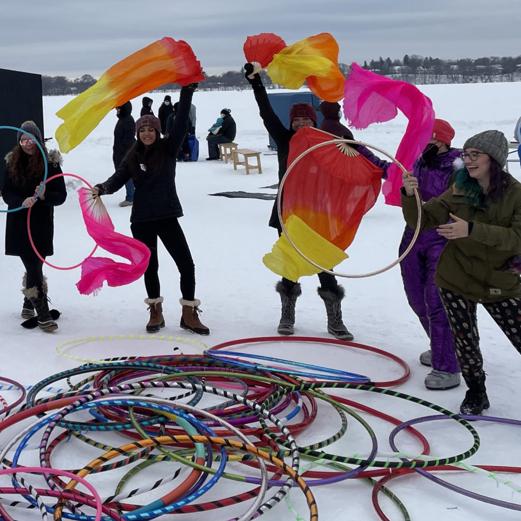 A group of performers wave colorful fabric in the air. A huge pile of hula hoops rests in front of them on the frozen lake.
